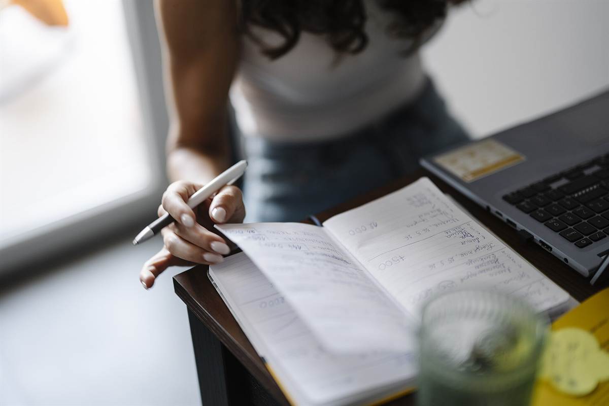 Gros plan sur une main qui prend des notes dans un carnet posé à côté d'un ordinateur portable