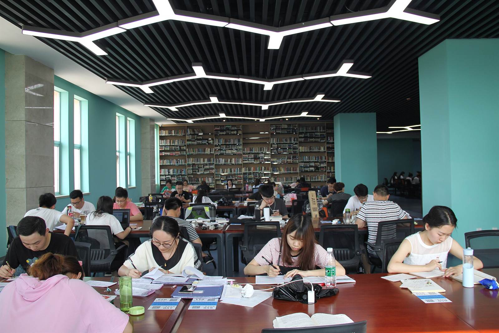 Grande salle de bibliothèque universitaire où des dizaines d'étudiantes et d'étudiants travaillent sur leurs cahiers, rayonnages de livres au fond