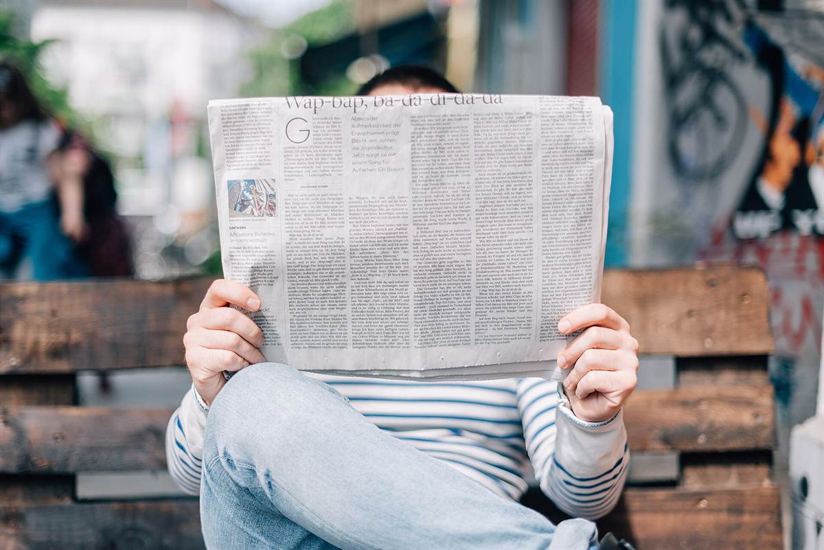 Personne assise sur un banc, le visage caché par le journal grand ouvert qu'elle est en train de lire